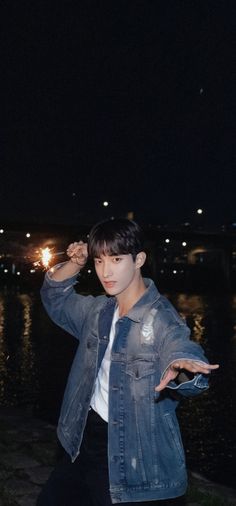 a young man standing next to a body of water wearing a jean jacket and white t - shirt