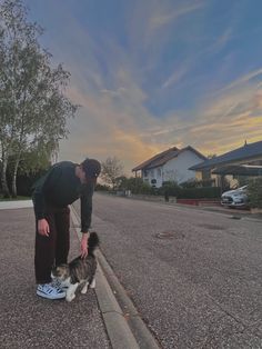 a man bending over to pet a dog on the side of the road at sunset