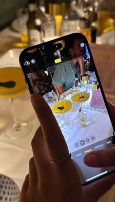 a person holding up a cell phone with wine glasses on the table in front of them