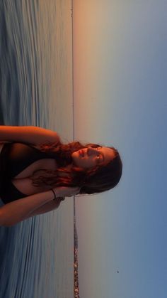 a woman in a black bathing suit standing next to the ocean with her arms behind her back