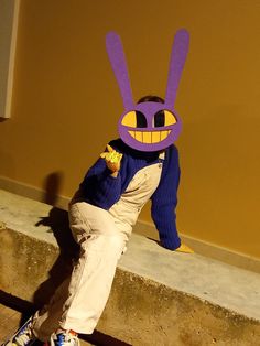 a young boy sitting on top of a cement wall wearing a purple bunny mask and smiling