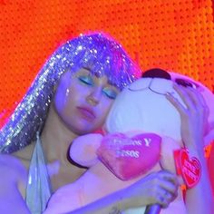 a woman with purple hair holding a white teddy bear in front of an orange background