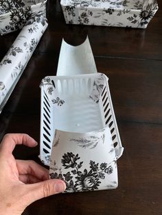 a person is holding some paper in front of two empty containers on a table with black and white flowers