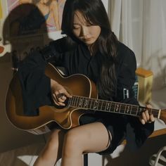 a woman sitting on a chair playing an acoustic guitar with her legs crossed and looking down