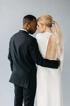 a man and woman standing next to each other in front of a gray wall wearing black suits