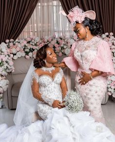 two women in wedding gowns sitting next to each other with flowers on the wall behind them