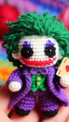 a small crocheted joker doll with green hair and purple pants holding a card