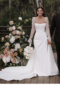 a bride and groom holding hands while standing on a wooden deck in front of flowers