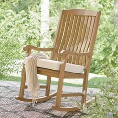 a wooden rocking chair sitting on top of a patio next to flowers and trees in the background