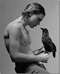 a shirtless man holding a bird in his right hand while sitting on a stool