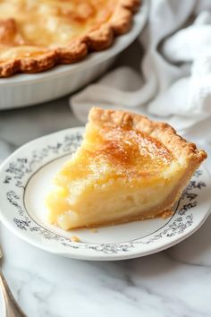 a slice of lemon pie on a plate next to a fork and bowl with another pie in the background