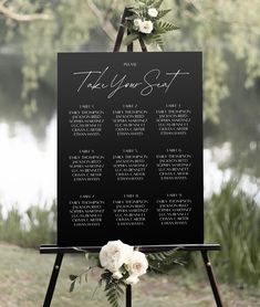 a black and white wedding seating sign with flowers on it in front of a lake