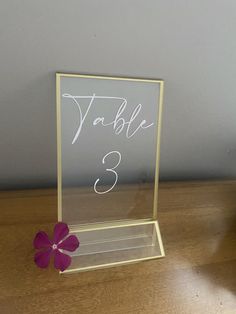 a table number holder with a purple flower on the side and a white sign that reads'table 3'in cursive writing