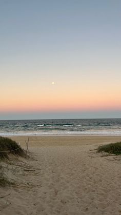 the sun is setting over the ocean and beach with grass in the foreground, as well as sand dunes on either side