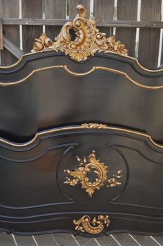 an ornate black and gold bed frame with intricate carvings on the headboard, in front of a wooden fence