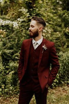 a man wearing a red suit and tie standing in front of some bushes with his hands in his pockets