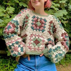 a girl with pink hair wearing a crocheted sweater in front of some bushes