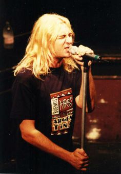 a man with long blonde hair holding a microphone in his right hand and looking at the camera