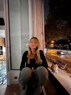 a woman sitting on a window sill in front of a building at night with her eyes closed