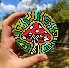 a hand holding up a colorfully painted wooden coaster in front of a house and trees