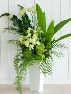 a white vase filled with lots of green flowers and greenery next to a wall