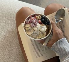 a person is reading a book and holding a bowl of cereal with fruit in it