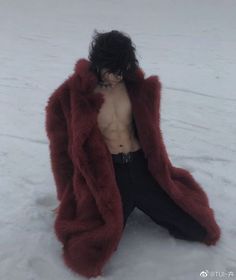 a shirtless man sitting in the snow wearing a red fur coat and black pants