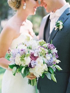 a bride and groom standing together in front of trees holding their wedding bouquet with purple and white flowers