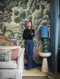 a woman standing next to a couch in front of a wall with trees on it