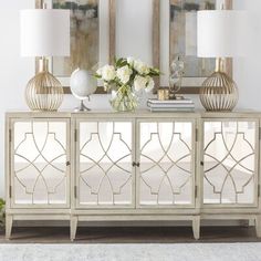 a white sideboard with two lamps and flowers on it in front of three mirrors