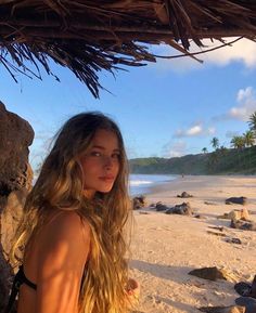 a beautiful young woman standing on top of a sandy beach next to the ocean under a thatched umbrella