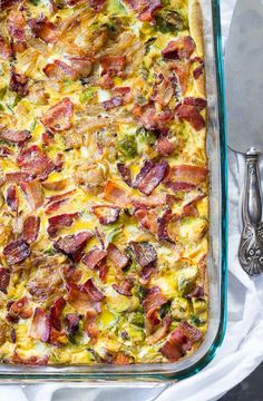 a casserole dish with bacon, broccoli and cheese in it next to two silver spoons