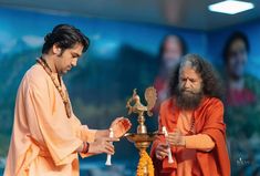 two men in orange robes holding candles and looking at each other while standing next to one another