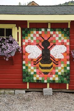 a quilt hanging on the side of a red building with flowers in front of it