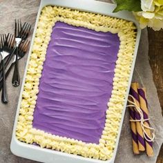 a square cake with purple frosting in a pan next to silverware and flowers