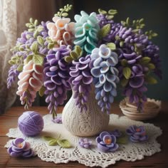 a white vase filled with purple flowers on top of a table next to balls of yarn