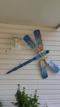 a blue and yellow dragonfly sculpture hanging on the side of a white house next to a potted plant