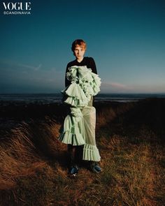 a woman standing on top of a grass covered field next to the ocean wearing a dress