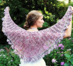 a woman holding up a pink shawl in front of some bushes and trees with purple flowers