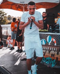 a man in white shirt and shorts standing on stage holding a microphone with other people behind him