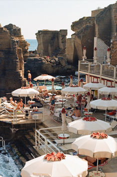 many people are sitting at tables near the water and some umbrellas on the beach