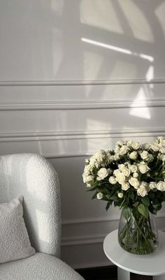 white roses in a vase on a table next to a chair