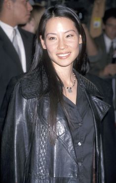 a woman with long hair wearing a black leather jacket and smiling at the camera while standing in front of a group of people