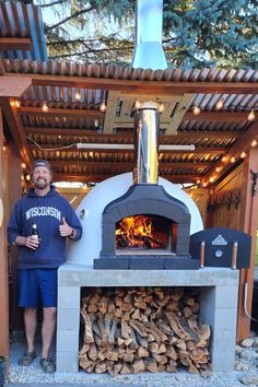 a man standing in front of an outdoor pizza oven with firewood stacked on it