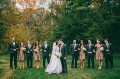 a bride and groom kissing in front of their wedding party outside on the grass with trees behind them