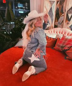 a woman sitting on top of a red bed wearing a cowboy hat and holding her hand up