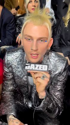 a man with blonde hair and piercings sitting in front of a crowd holding a microphone