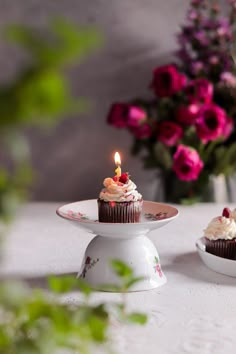 two cupcakes sitting on top of a cake plate with a lit candle in the middle