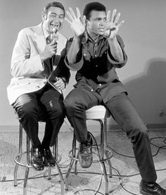 two men sitting on stools with their hands in the air and one holding up his hand