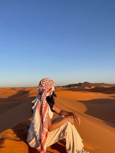 a person sitting on top of a sand dune in the desert wearing a headdress